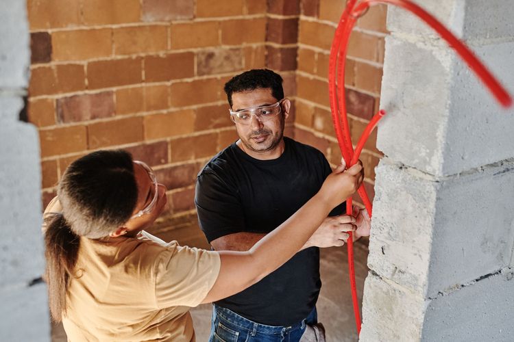 Two people in safety glasses guiding red plastic water pipe along a brick wall on a building site