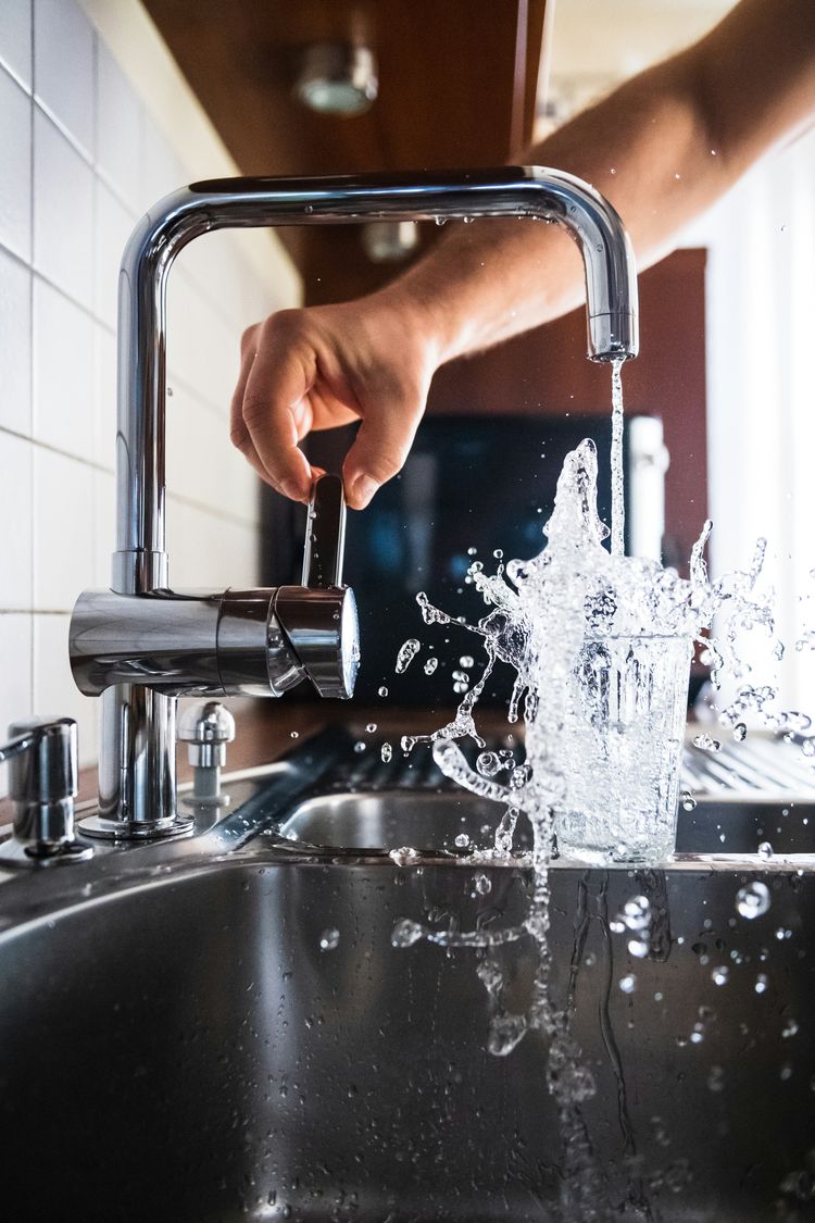 Hand opening a chrome kitchen tap with water splashing into a glass