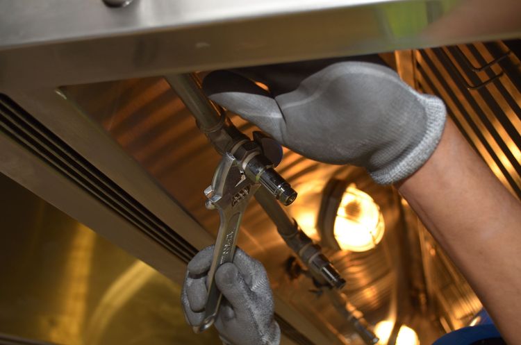 Gloved hands tightening a pipe fitting with a spanner under a stainless steel extractor hood